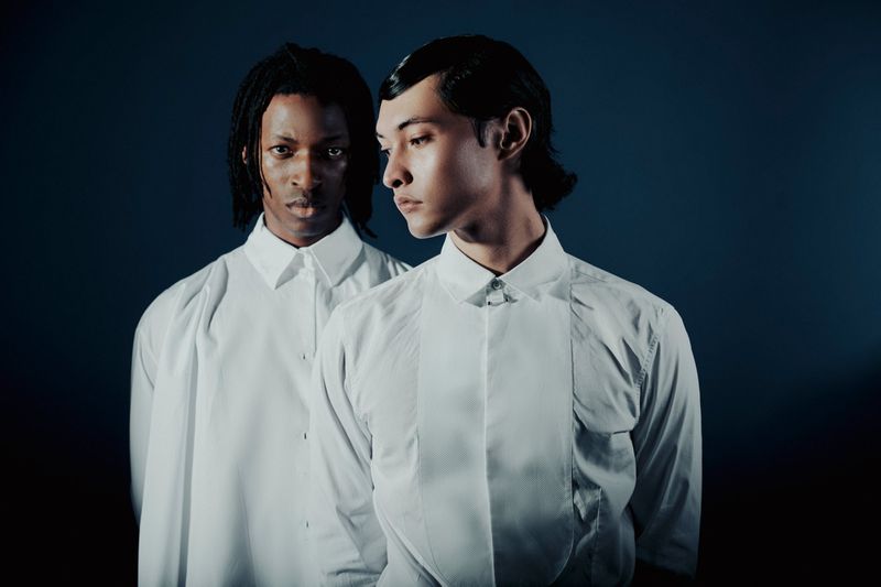 Two young adult men with clean skin and styled dark hair pose closely in white collared shirts against a dark blue studio backdrop.