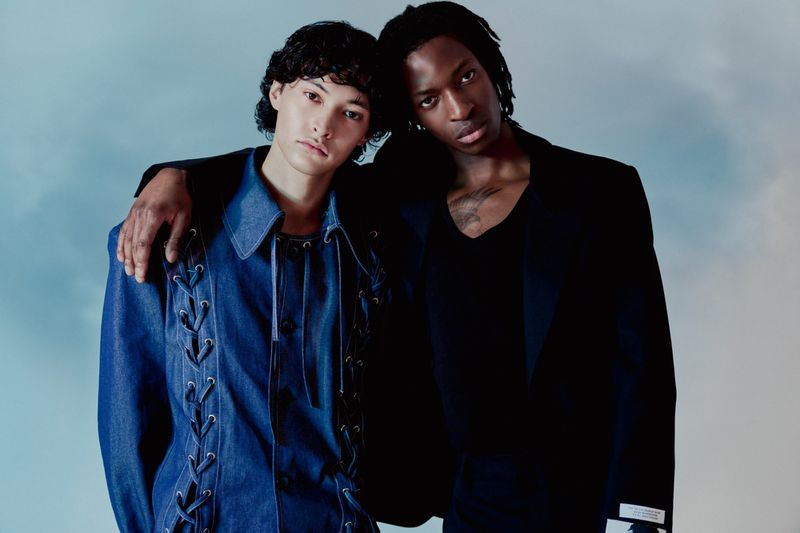 Two men pose in a studio with a cloudy sky background. One wears a blue denim shirt with lace-up detailing, the other a black blazer.