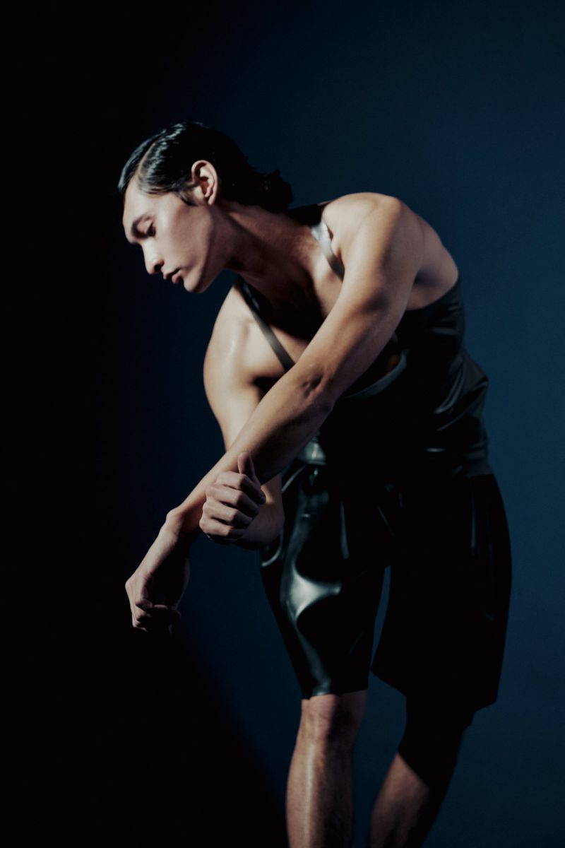 Muscular adult male in wet-look tank top and shorts poses in a dimly lit, dark environment with slicked-back hair.