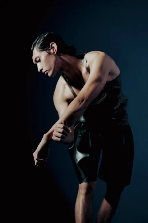 Muscular adult male in wet-look tank top and shorts poses in a dimly lit, dark environment with slicked-back hair.
