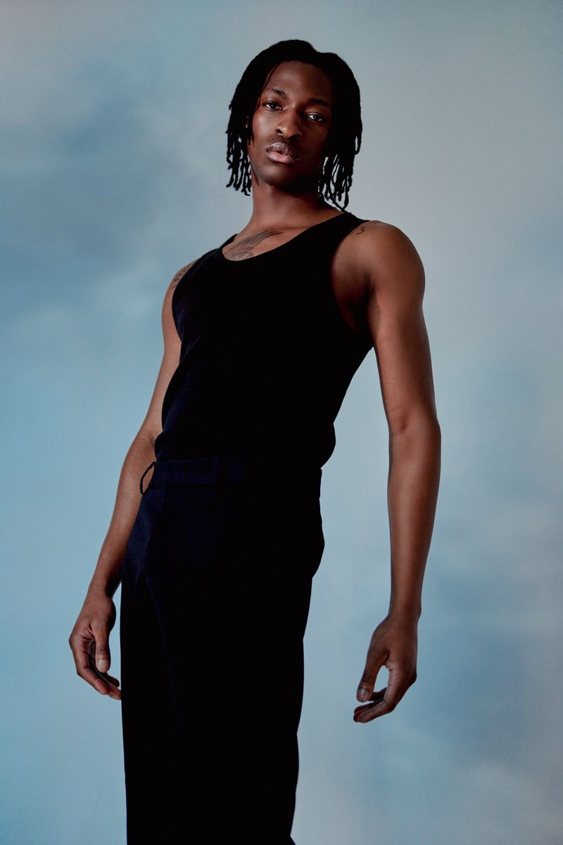 Studio portrait of a young Black male model in a tank top and pants against a blue backdrop. Fashion editorial and lifestyle imagery.