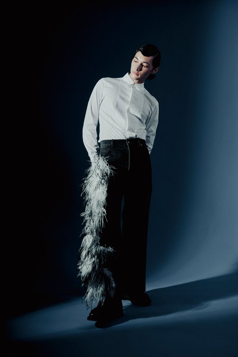 Full-length shot of a young adult wearing a white button-down, black pants with a feathered accent, and dark shoes, standing in a studio.