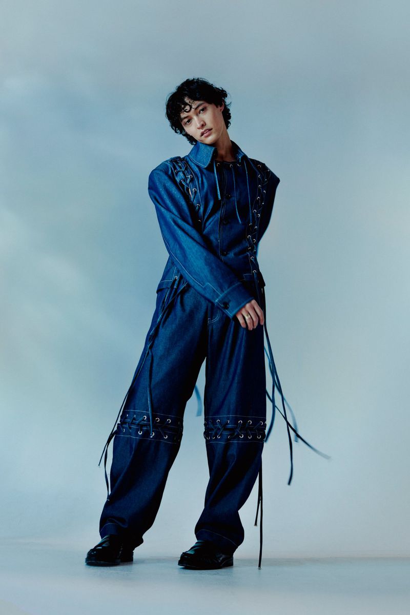 Young adult woman with short dark hair poses in an indoor studio wearing a denim jacket and wide leg pants embellished with grommets.