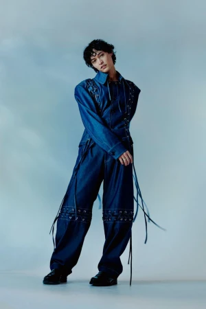 Young adult woman with short dark hair poses in an indoor studio wearing a denim jacket and wide leg pants embellished with grommets.