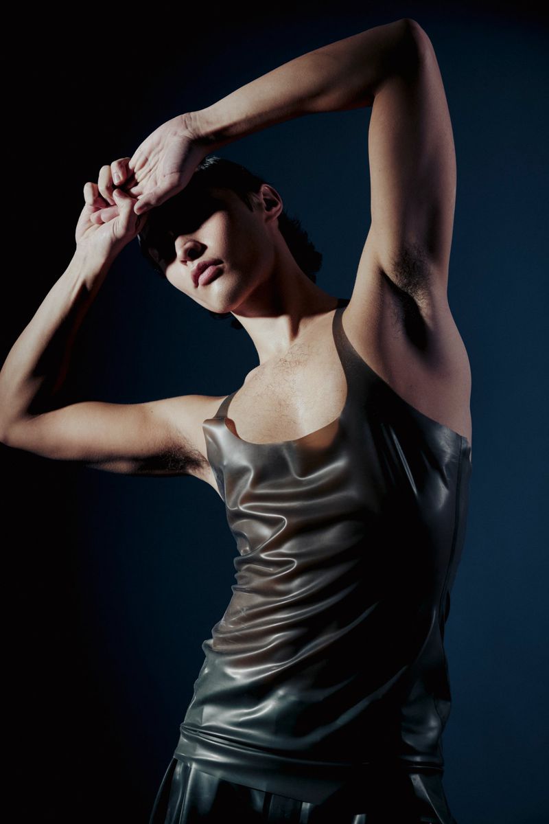 Fashionable adult model poses in a black latex tank top and skirt in a studio, hands raised over head against a dark background.