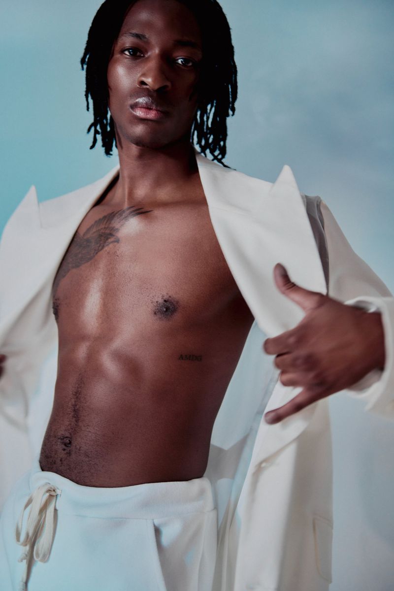 Portrait of a Black man with dreadlocks, wearing a white jacket and pants, showcasing his torso against a soft blue background.