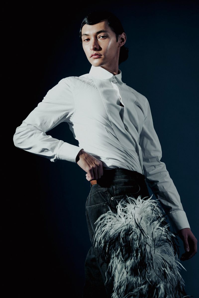Fashion portrait of a slender adult with dark hair wearing an unbuttoned white shirt, jeans, and feathered chaps on a dark background.