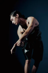 Dancer in a dark tank top and shorts posing dramatically against a blue studio background.