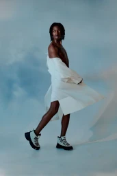 Black man with locs wearing a white open jacket and hiking boots against a cloudy sky background