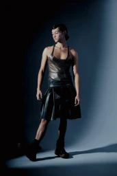 Model in a metallic tank top and dark pleated shorts standing against a grey studio background