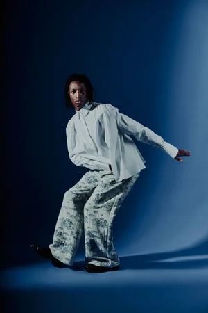 Black man in a white button-down shirt and patterned trousers posing against a deep blue gradient backdrop.