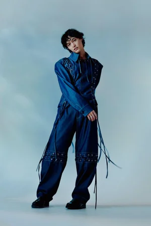 A man in a blue denim outfit with laced details posing against a soft gradient background for The Rakish Gent