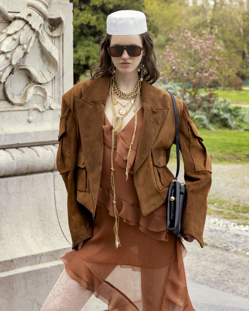 A young woman with long brown hair poses outdoors wearing a brown jacket, a light brown dress, and a white kufi cap near a stone wall.