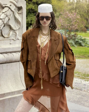 A young woman with long brown hair poses outdoors wearing a brown jacket, a light brown dress, and a white kufi cap near a stone wall.