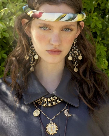 Close-up of a woman with brown hair, blue eyes, wearing dangling earrings, a sun pendant necklace, and a patterned scarf headband.