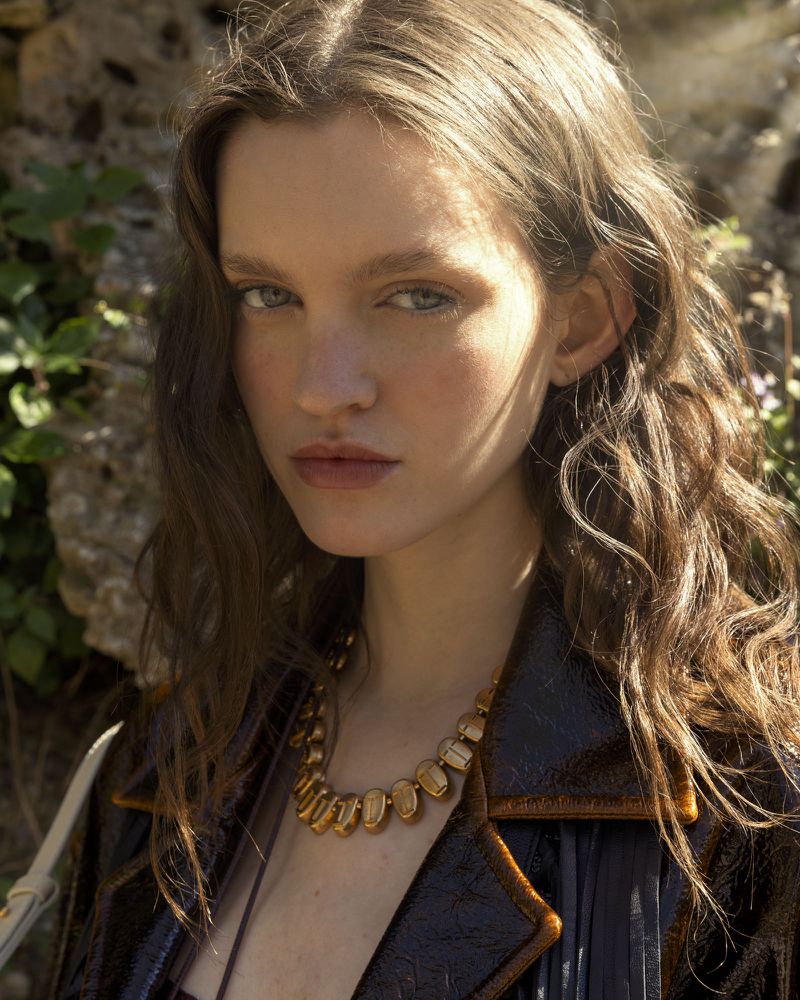 Portrait of a young woman with wavy brown hair, wearing a brown jacket and a gold necklace, posing against a natural stone backdrop.