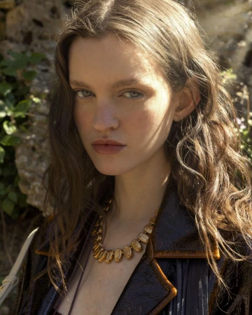 Portrait of a young woman with wavy brown hair, wearing a brown jacket and a gold necklace, posing against a natural stone backdrop.