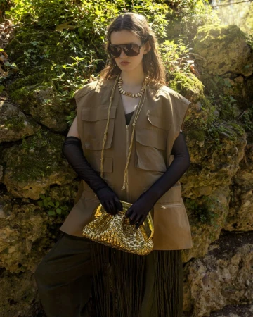 A woman with long brown hair poses in a vest, long gloves and sunglasses, holding a gold clutch in a natural, outdoor setting.