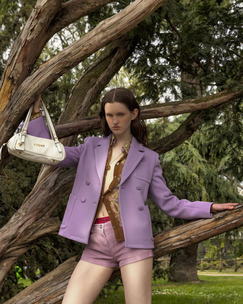 A woman wearing a purple blazer and shorts, holding a white purse with the Versace logo, stands in front of a tree in a park.
