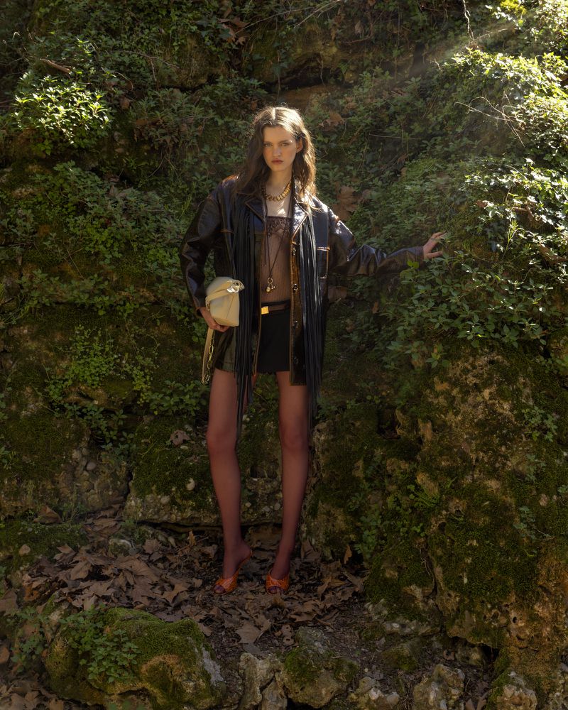 An adult woman poses in a forest, wearing a fringed jacket, short skirt, sheer top, hose, and orange heels, carrying a cream-colored handbag.
