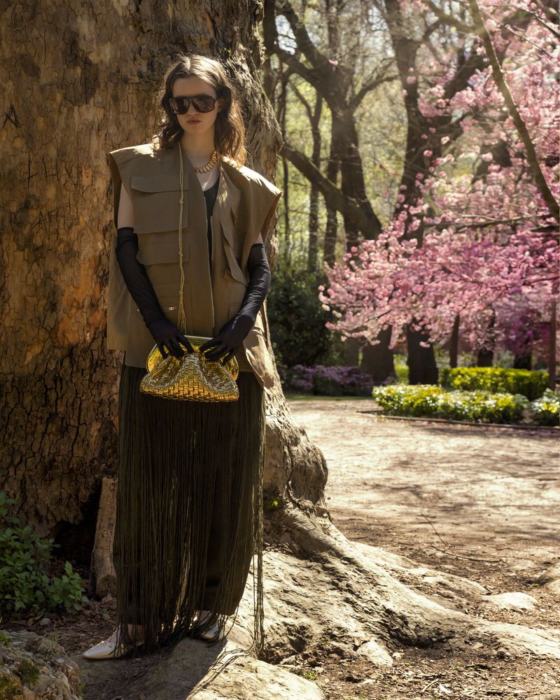 Woman in vest and sunglasses holding a purse in an outdoor setting with cherry blossom trees and a large tree trunk. Fashion editorial.