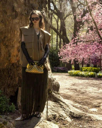 Woman in vest and sunglasses holding a purse in an outdoor setting with cherry blossom trees and a large tree trunk. Fashion editorial.