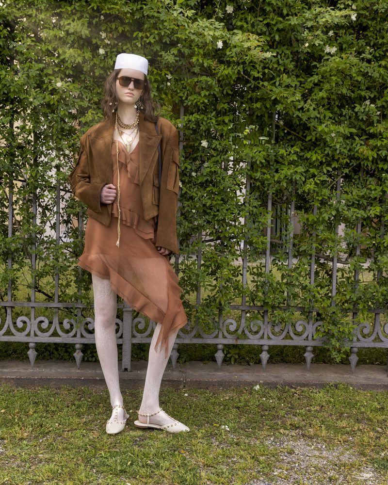A woman in a brown dress and jacket poses outdoors in front of greenery, wearing sunglasses, a white hat, and flats. Fashion editorial.