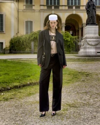 Model in an oversized black blazer and matching trousers with a white cap standing outdoors near a historic building