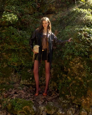 Model standing outdoors in front of a mossy rock wall wearing a dark fringed leather jacket and holding a cream handbag
