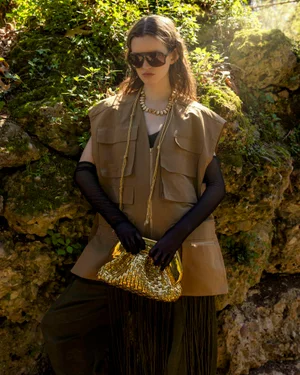 Model wearing dark sunglasses, utilitarian brown vest, black gloves, and holding a gold clutch in a natural woodland setting