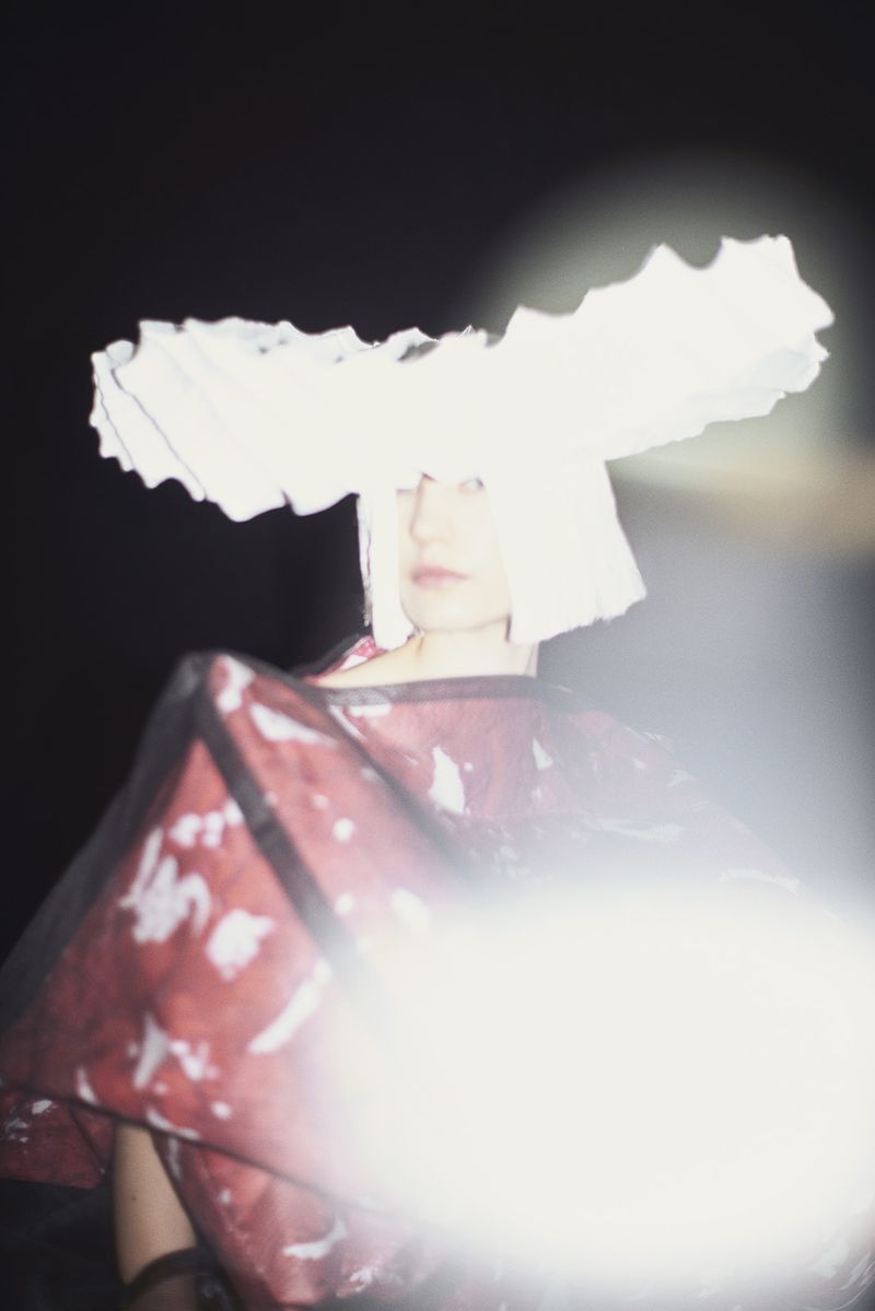 A woman with short white hair poses wearing a large white hat and patterned red top against a dark background with bright lighting.