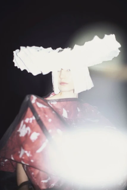 A woman with short white hair poses wearing a large white hat and patterned red top against a dark background with bright lighting.