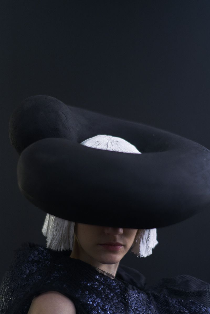 Woman wearing a black sculptural hat and a sequined top against a dark background in a studio setting, fashion-forward and artistic.