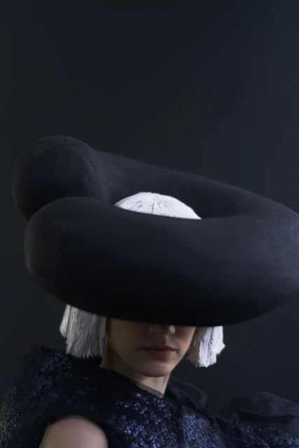 Woman wearing a black sculptural hat and a sequined top against a dark background in a studio setting, fashion-forward and artistic.