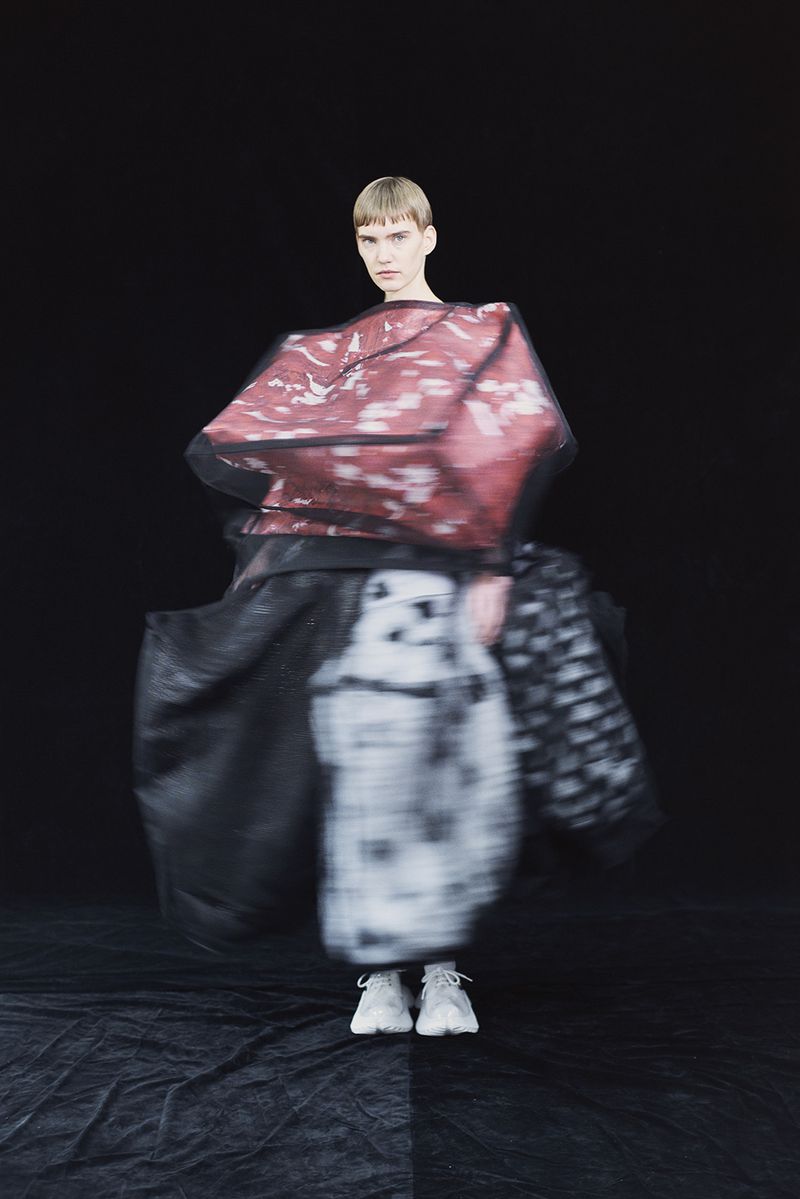 Fashion model strikes pose in avant-garde outfit in photo studio, featuring abstract patterns and unique design elements against black backdrop.