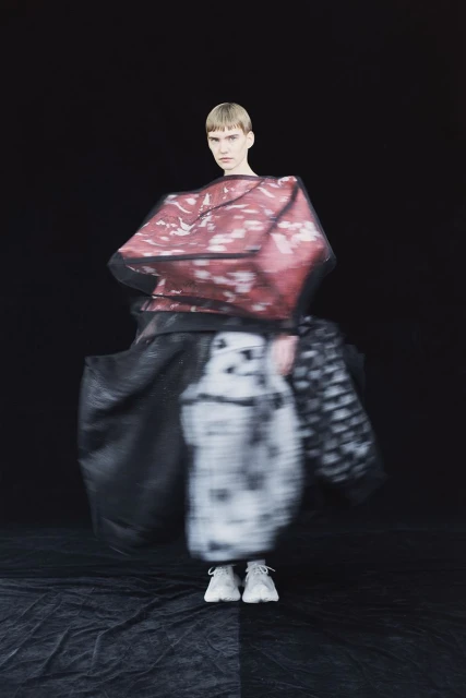 Fashion model strikes pose in avant-garde outfit in photo studio, featuring abstract patterns and unique design elements against black backdrop.