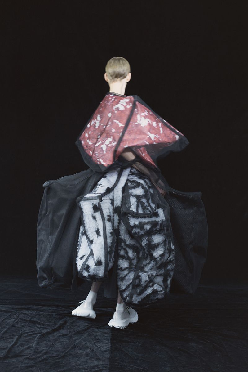 Fashion model in a black, red, and white garment, posing on a black and grey floor against a black background in a studio shot.