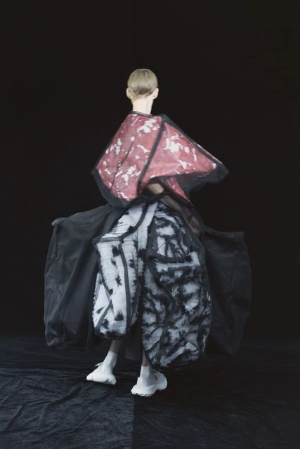 Fashion model in a black, red, and white garment, posing on a black and grey floor against a black background in a studio shot.
