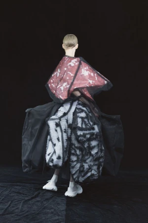 Fashion model in a black, red, and white garment, posing on a black and grey floor against a black background in a studio shot.