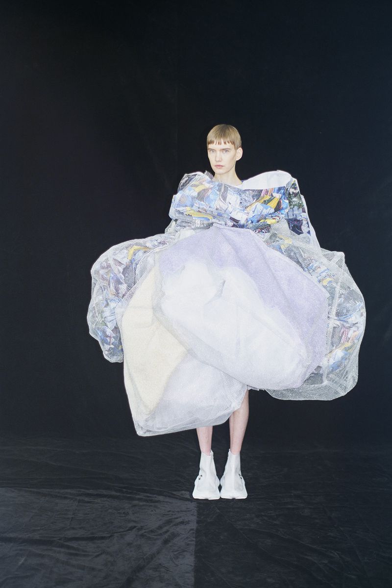 Fashion photo of a young woman with short blond hair in a puffy, white, translucent dress with white platform boots against a dark background.