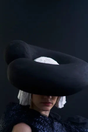 Model wearing a large sculptural black hat and dark textured top against a dark background