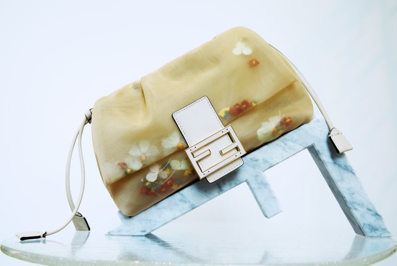 Beige sheer handbag with floral details, featuring the Fendi logo, placed on a marble-patterned Fendi logo stand, against a white backdrop.