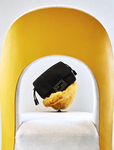 Black Fendi purse on a yellow furry stand, placed on a white chair with a yellow frame in a brightly lit indoor setting.