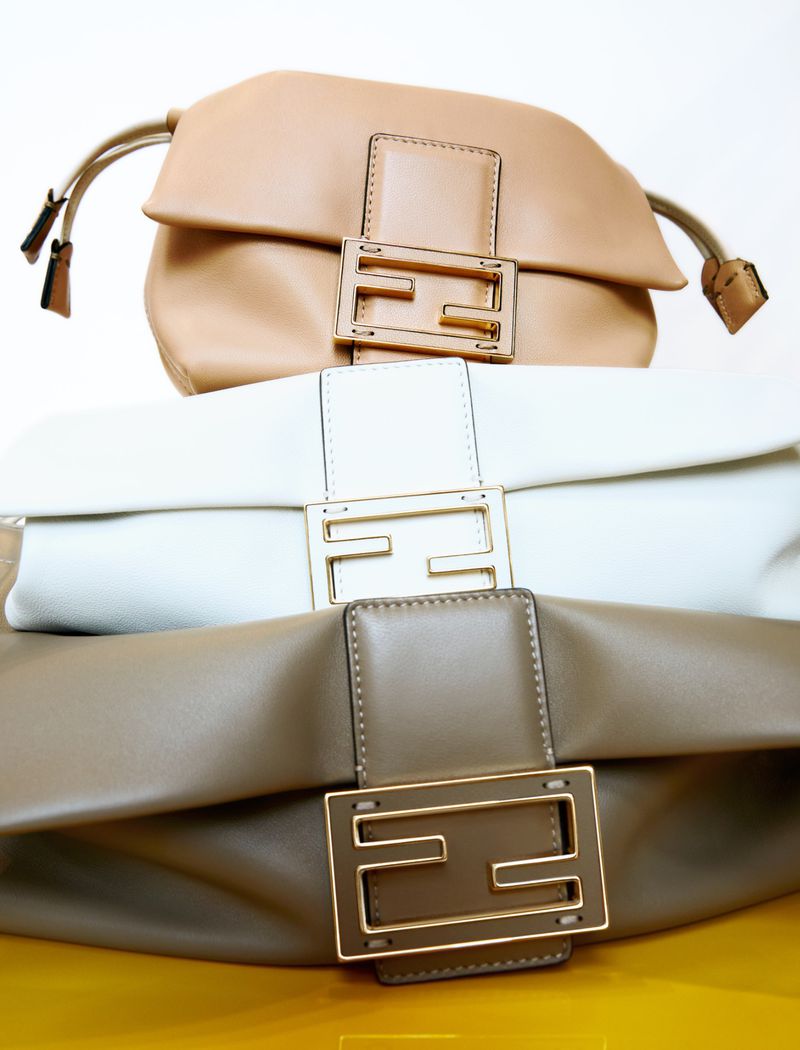 Three Fendi purses are stacked vertically against a yellow background. The bags are cream, white, and beige and feature a metal Fendi logo.