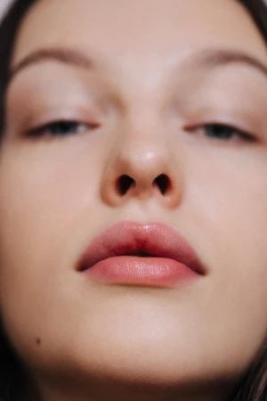 Close-up portrait of an adult woman with fair skin, light pink lipstick, and dark hair, set against a plain, neutral background.