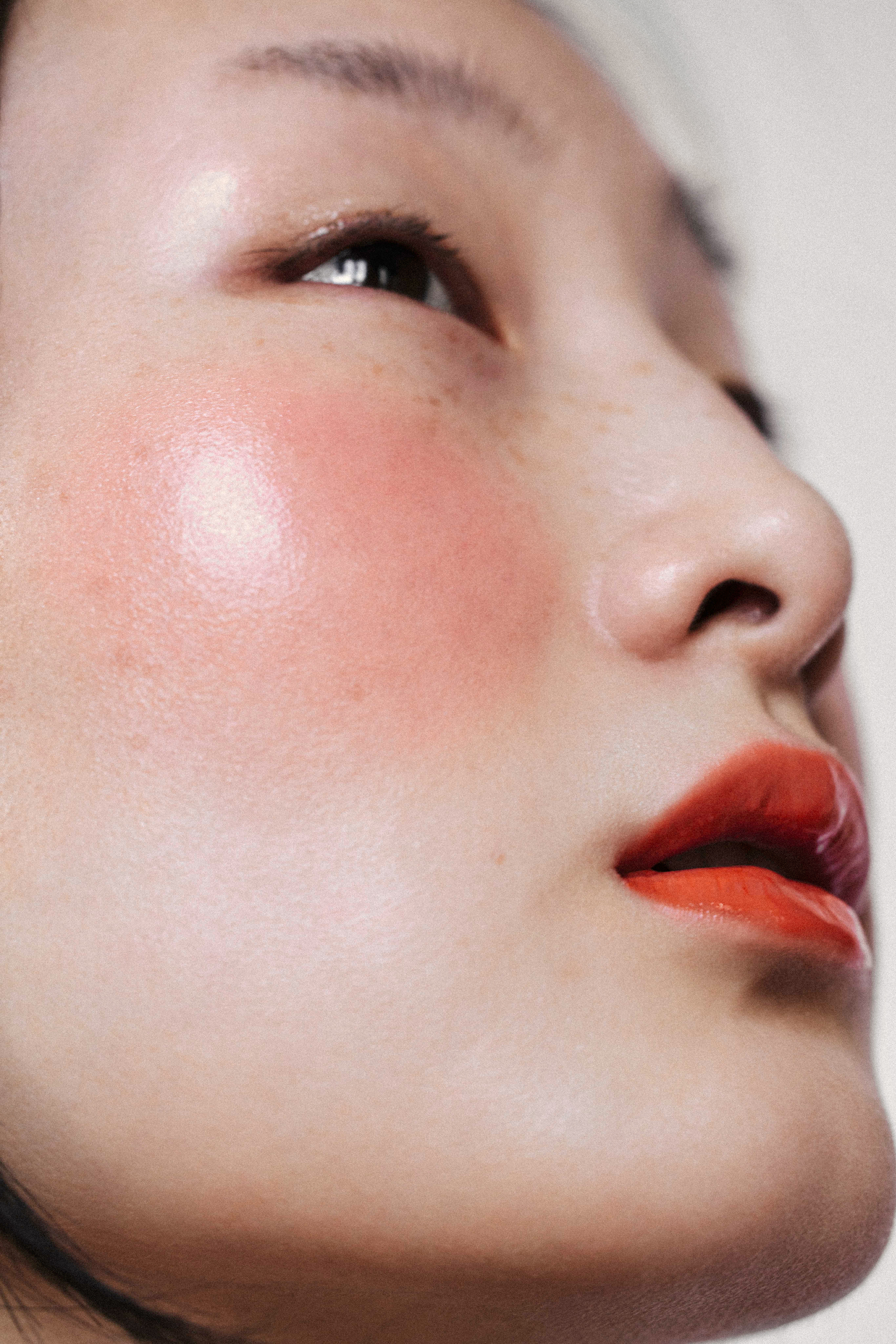 Close-up portrait of an Asian woman with glossy red lipstick and pink blush, looking up with a soft, glowing complexion in natural light.