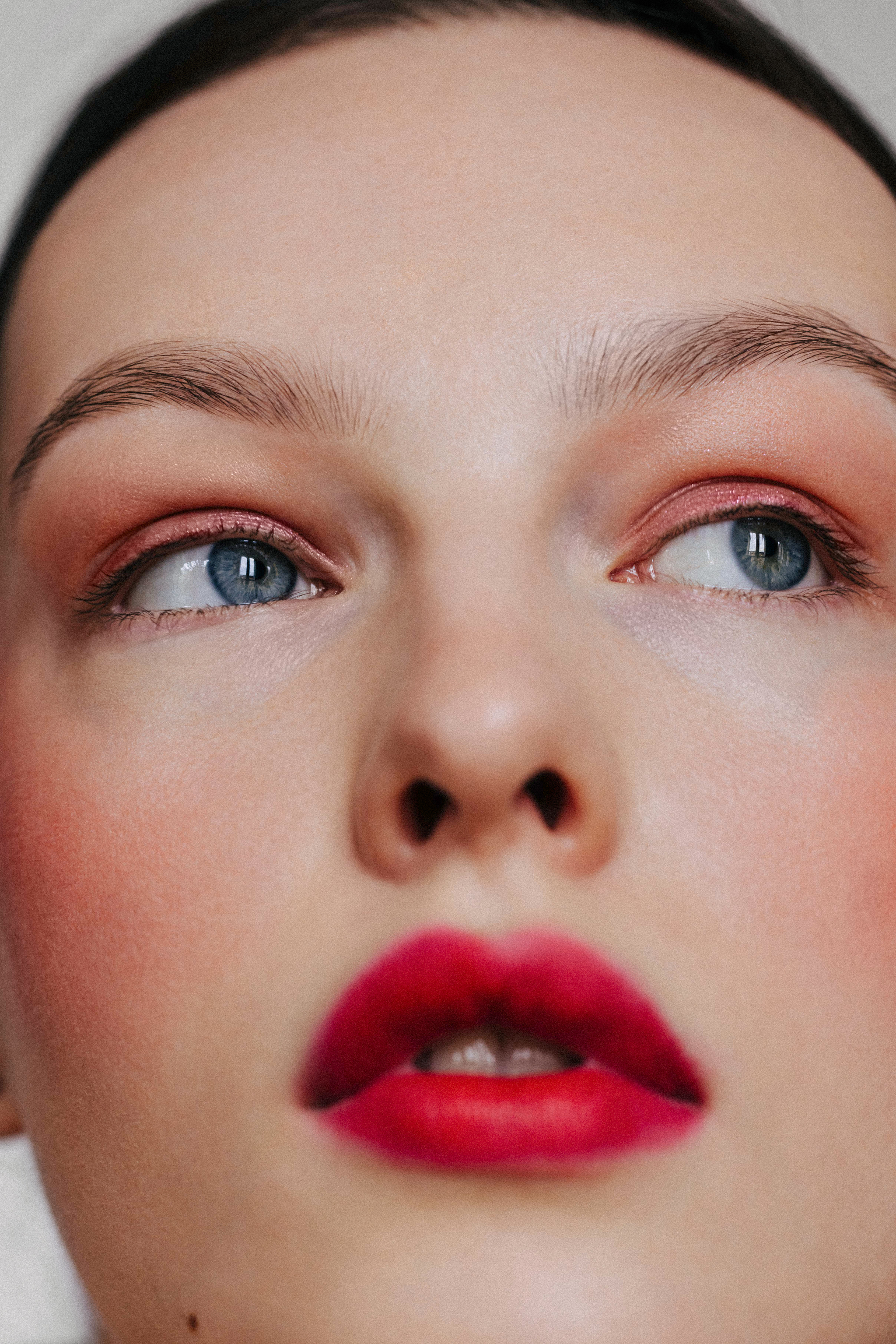 Extreme close-up of a woman's face with bold red lipstick, soft peach eyeshadow, and blue eyes looking upward.