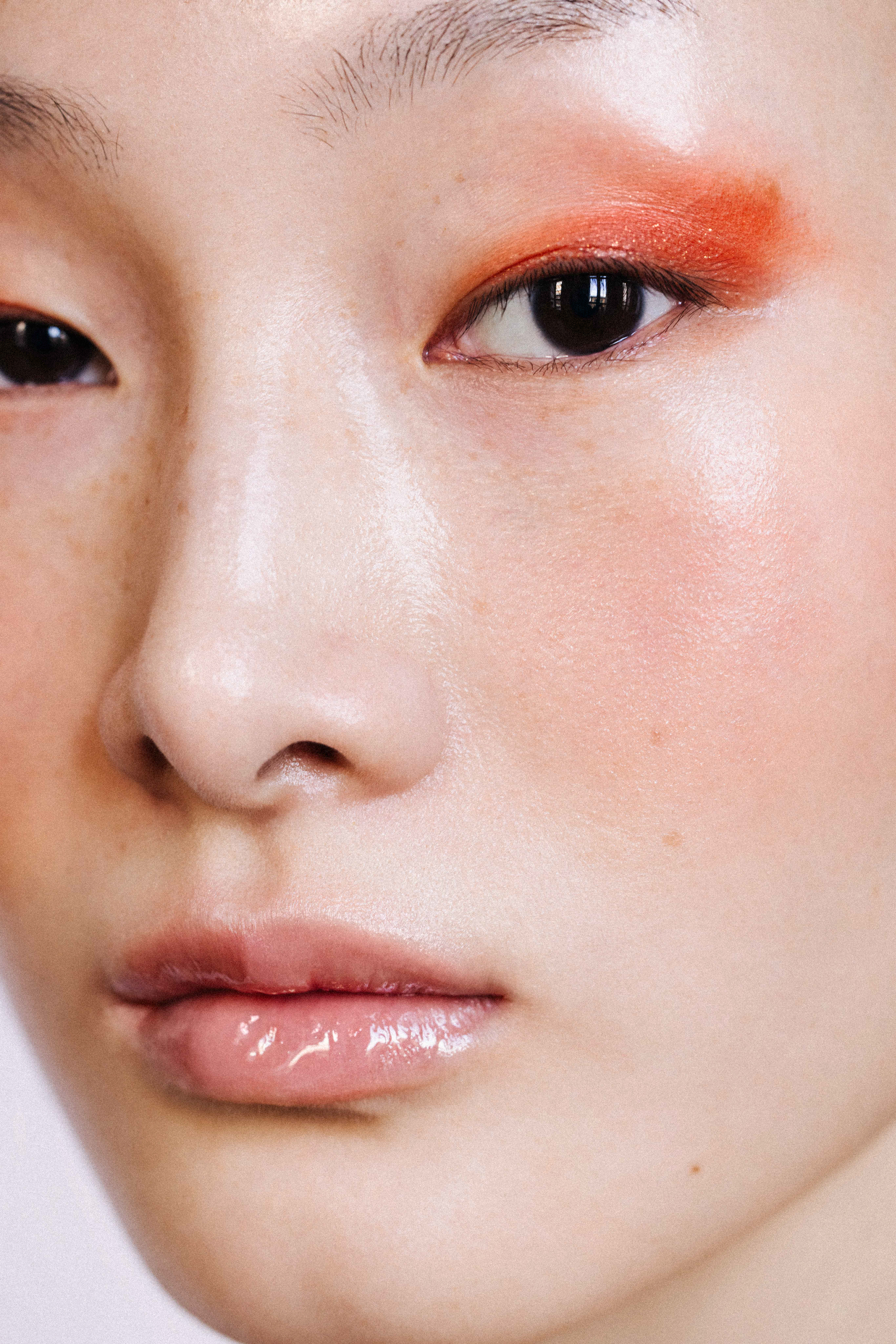 Close-up beauty portrait of a model with vibrant orange eyeshadow and glossy lips for Lucie, retouched by Ruslana Kharchenko.