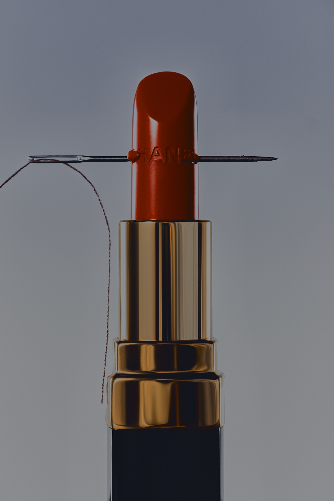 Close-up of a red lipstick with a sewing needle stuck through it, set against a soft grey background, beauty shot.