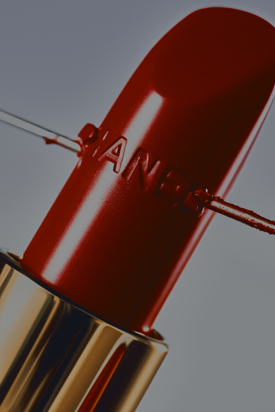 Close-up of a red Chanel lipstick with a gold tube, set against a muted grey background, highlighting the product's design and color.